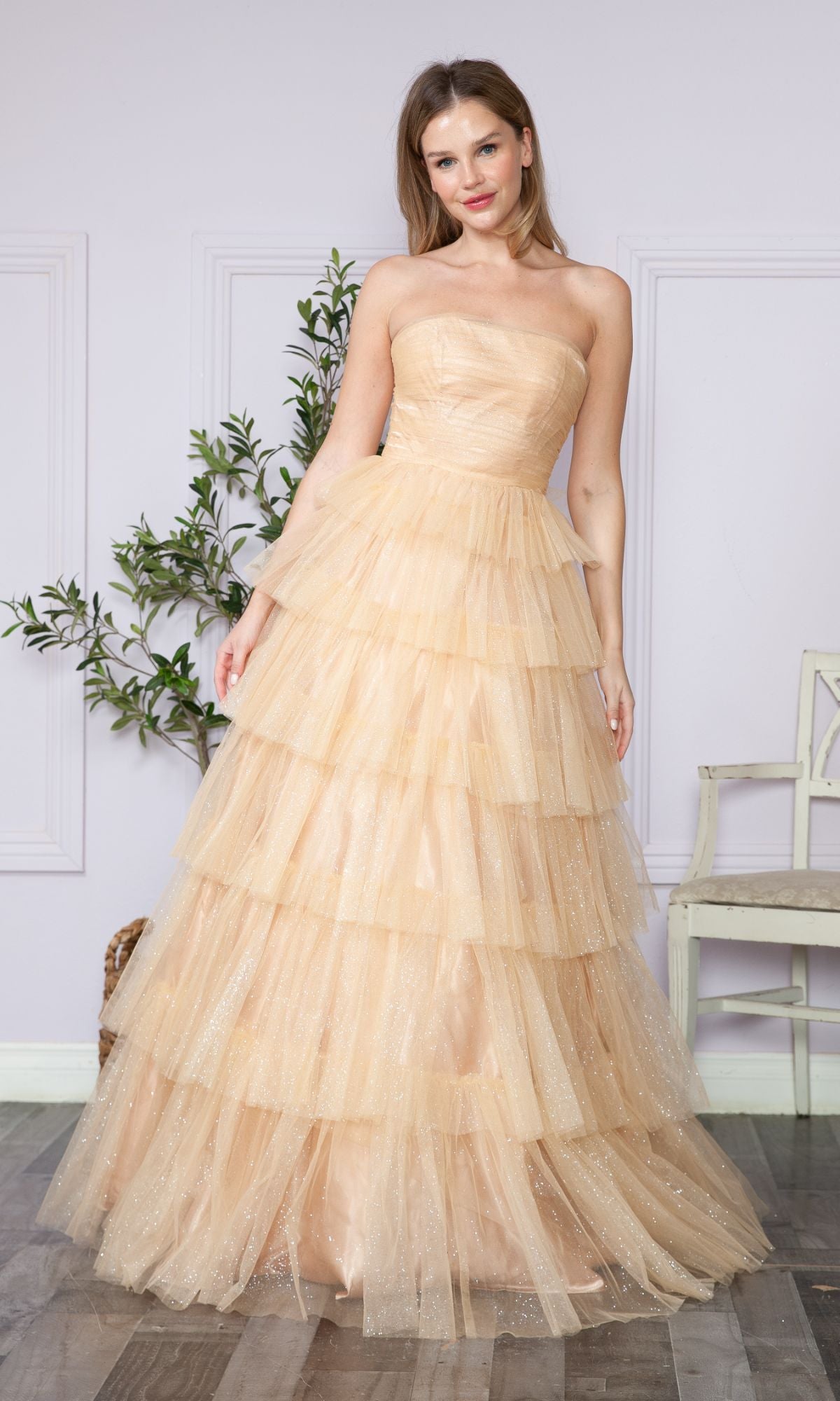 A woman in the Strapless Long Tiered Glitter Prom Ball Gown 9386, crafted in layered champagne tulle, stands indoors beside a white chair and a plant.